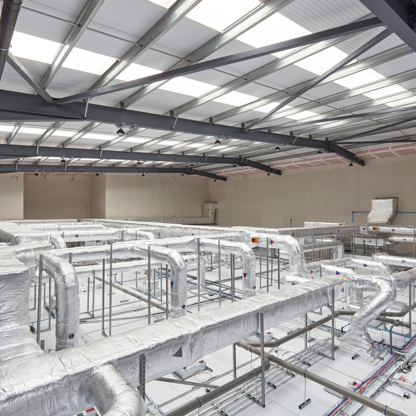 Large industrial interior featuring a complex HVAC duct system wrapped in insulation, supported by metal framing. A worker is inspecting the installation beneath a steel-framed roof with translucent ceiling panels.