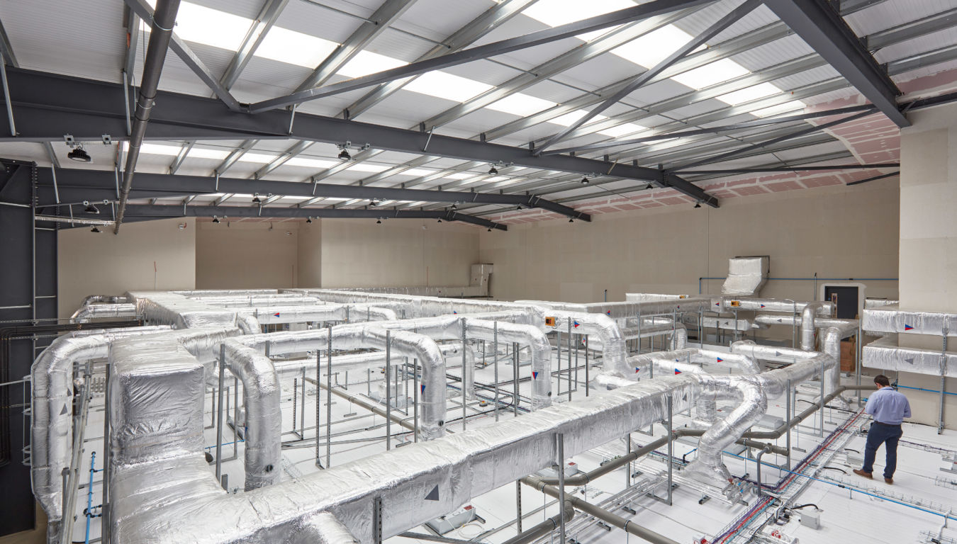 Large industrial interior featuring a complex HVAC duct system wrapped in insulation, supported by metal framing. A worker is inspecting the installation beneath a steel-framed roof with translucent ceiling panels.
