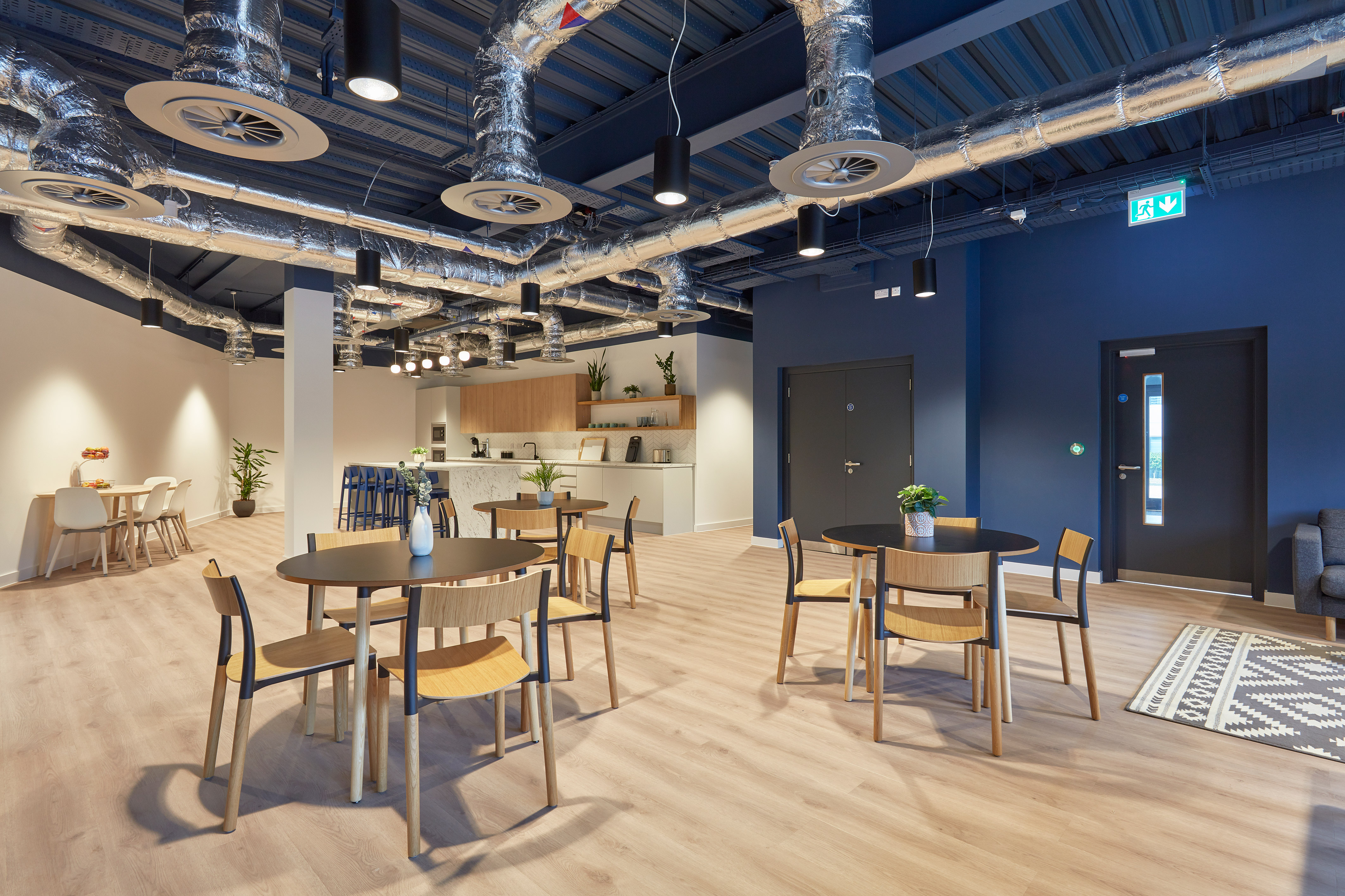 Modern office break room with round tables, wooden chairs, and a kitchenette. Exposed ductwork on the ceiling, minimalistic decor, warm lighting