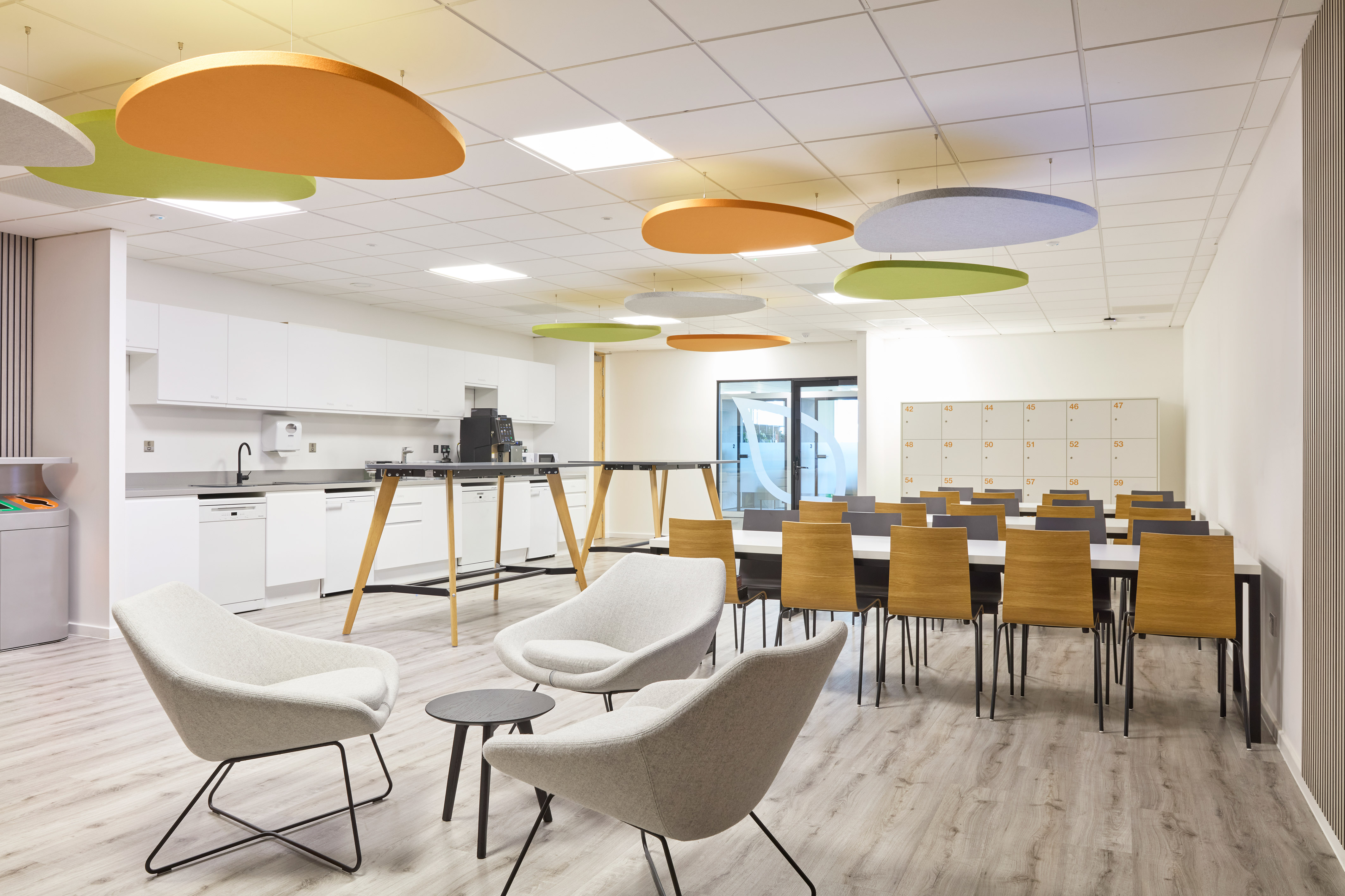 Modern office break room with white kitchen, long dining tables, and wooden chairs. Colorful ceiling panels and soft seating create a welcoming atmosphere.