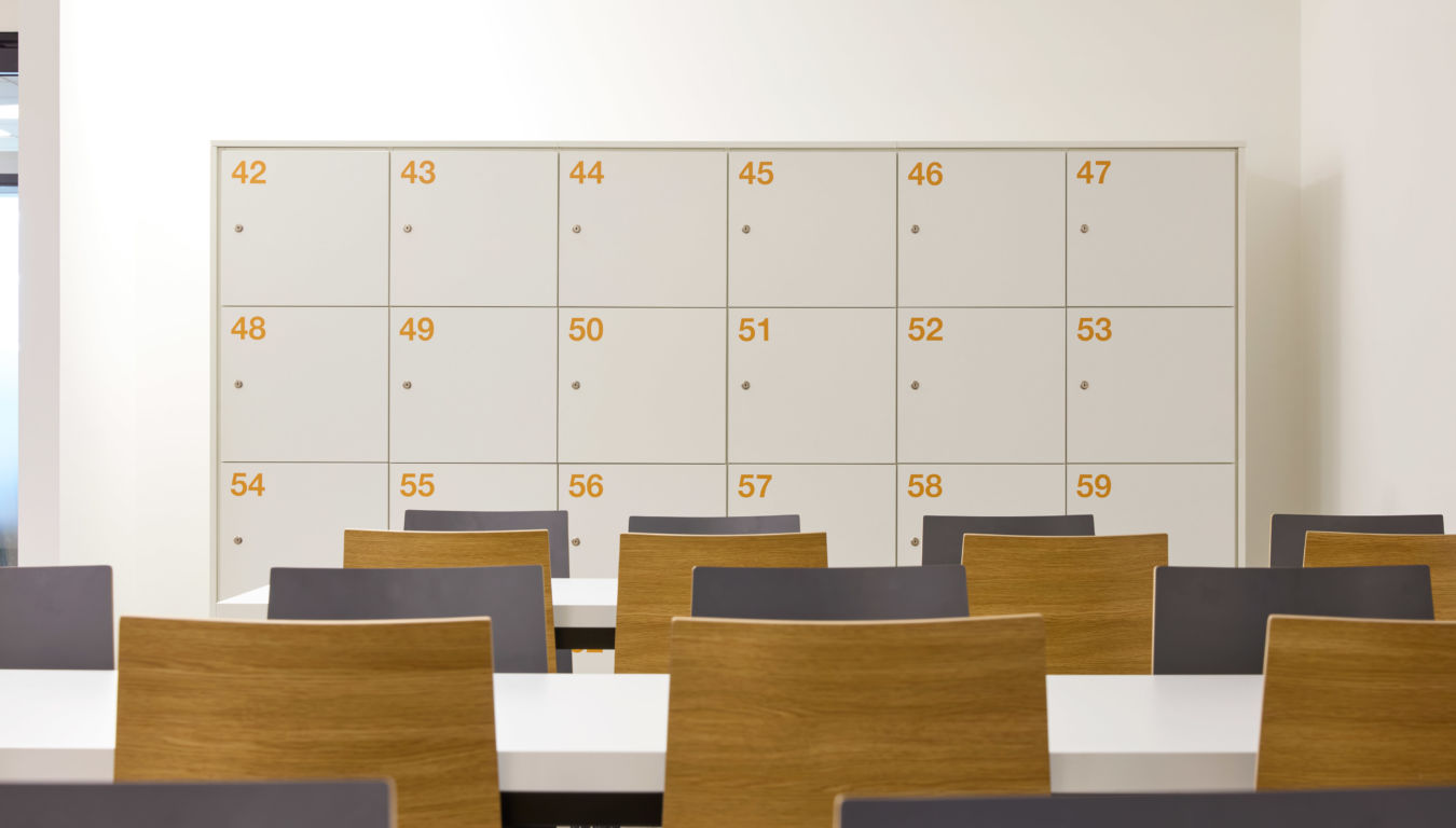 modern classroom features numbered white lockers against a white wall. In the foreground, there are white tables and chairs with wooden backs. The atmosphere is clean and organized