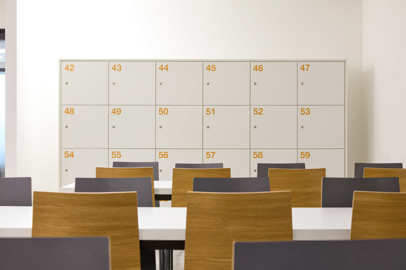 modern classroom features numbered white lockers against a white wall. In the foreground, there are white tables and chairs with wooden backs. The atmosphere is clean and organized