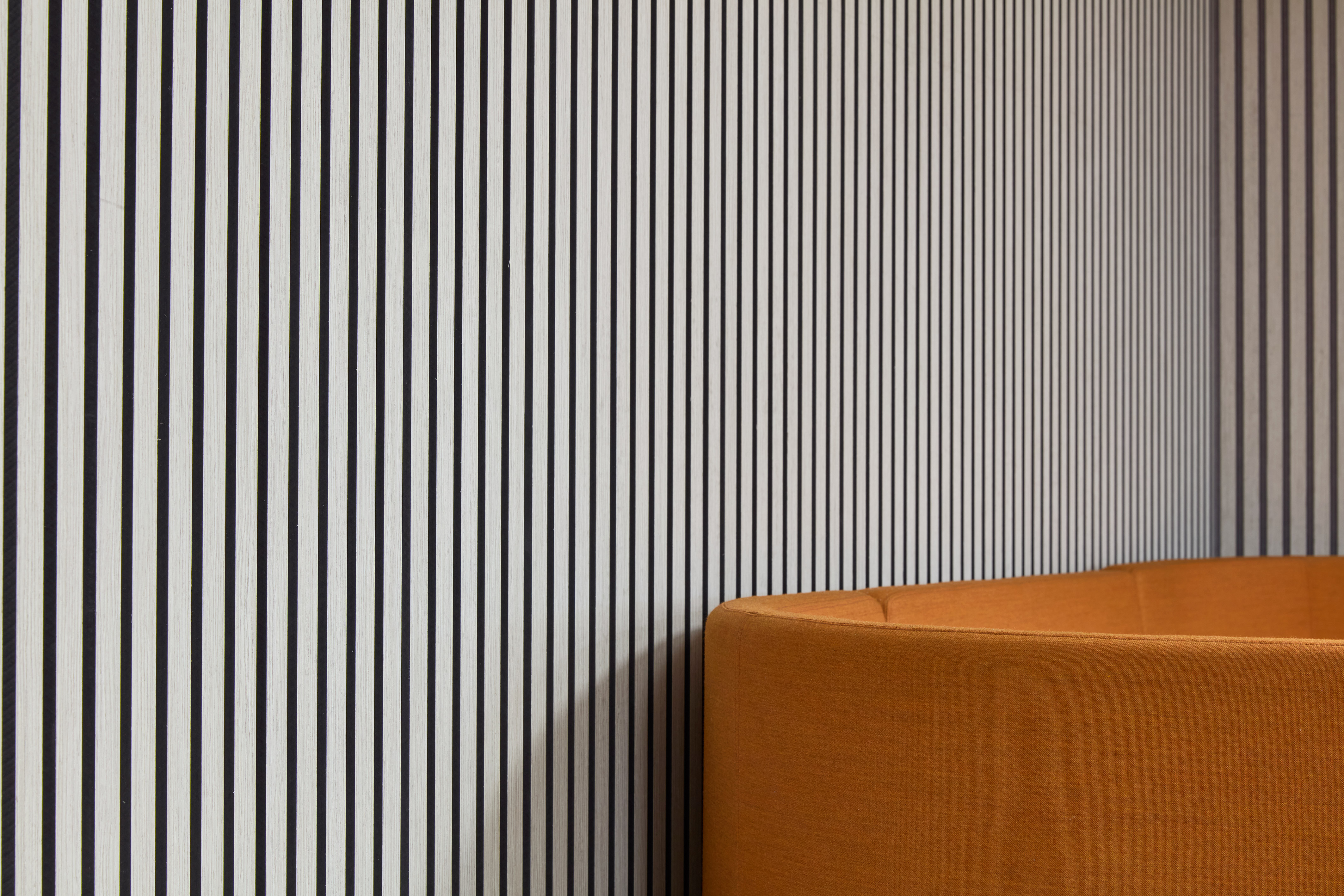 Orange upholstered couch curves against a wall with black and white vertical stripes, creating a modern, contrasting interior design setting.