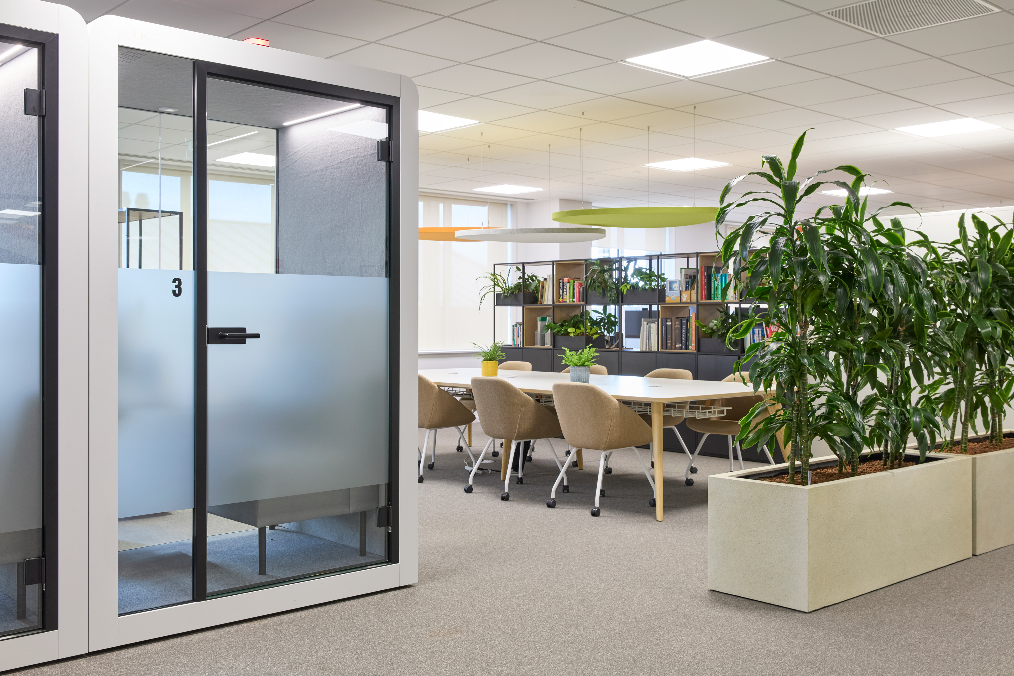 Modern open office with a private phone booth, collaborative meeting table, neutral chairs, and various indoor plants.