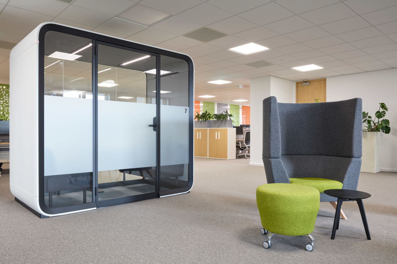 Modern office space with a sleek glass meeting pod on the left and a cozy seating area on the right, featuring a gray chair, green ottoman, and small black table