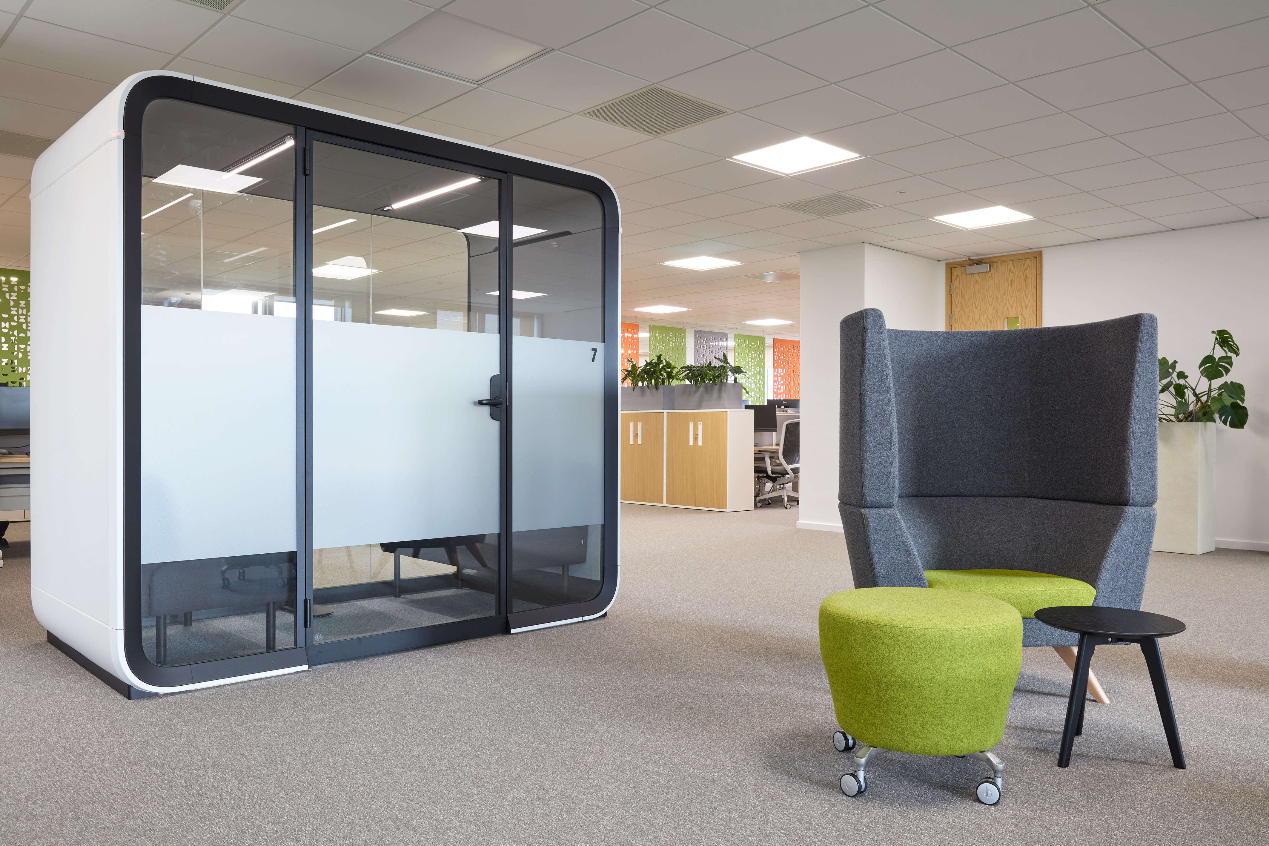 Modern office space with a sleek glass meeting pod on the left and a cozy seating area on the right, featuring a gray chair, green ottoman, and small black table