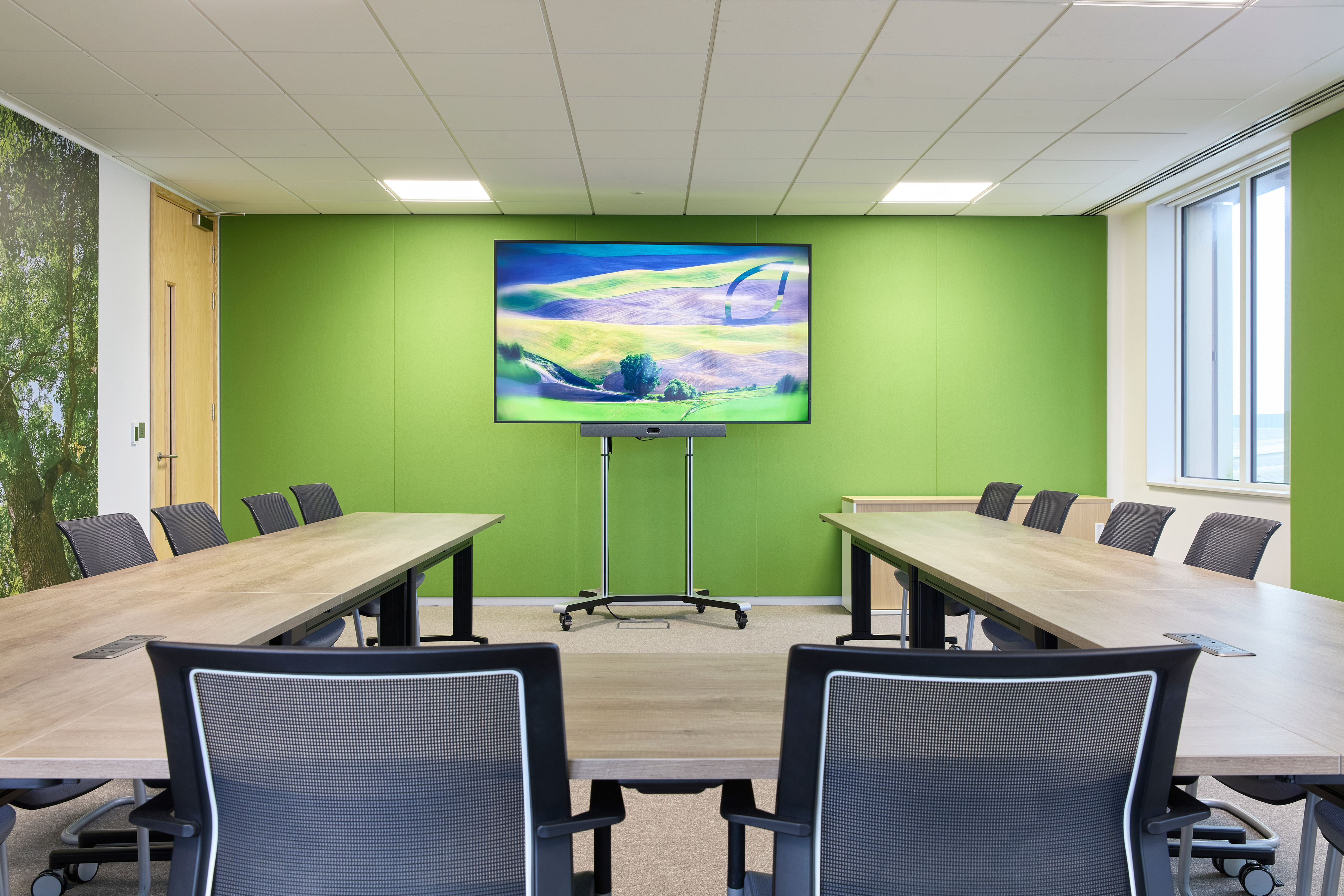 Conference room featuring a long wooden table surrounded by office chairs, a large screen displaying scenic countryside imagery, set against vibrant green walls and natural lighting from windows.