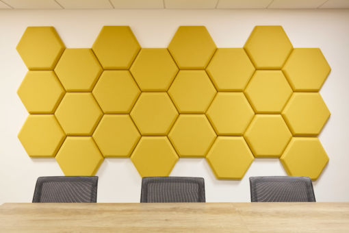 Yellow hexagonal acoustic panels are arranged in a honeycomb pattern on a white wall above a wooden conference table with three gray mesh chairs in the foreground.
