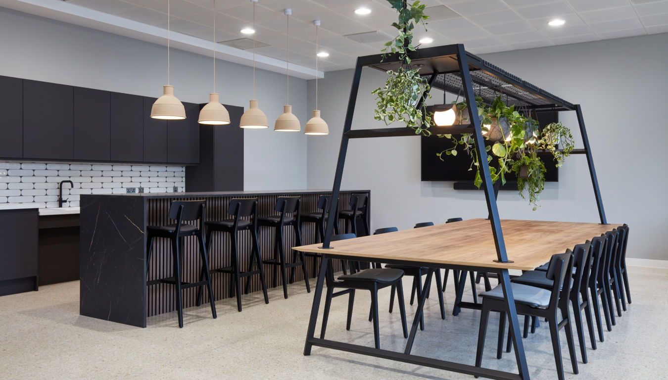 Large wooden table with black chairs placed under a metal frame with hanging plants, situated in a modern kitchen featuring a black counter, pendant lights, and white brick backsplash.