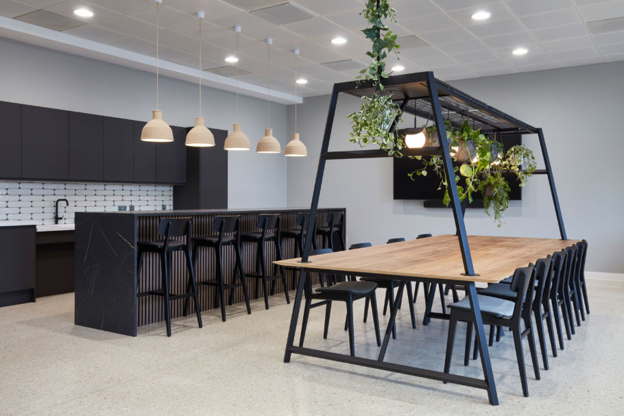Large wooden table with black chairs placed under a metal frame with hanging plants, situated in a modern kitchen featuring a black counter, pendant lights, and white brick backsplash.