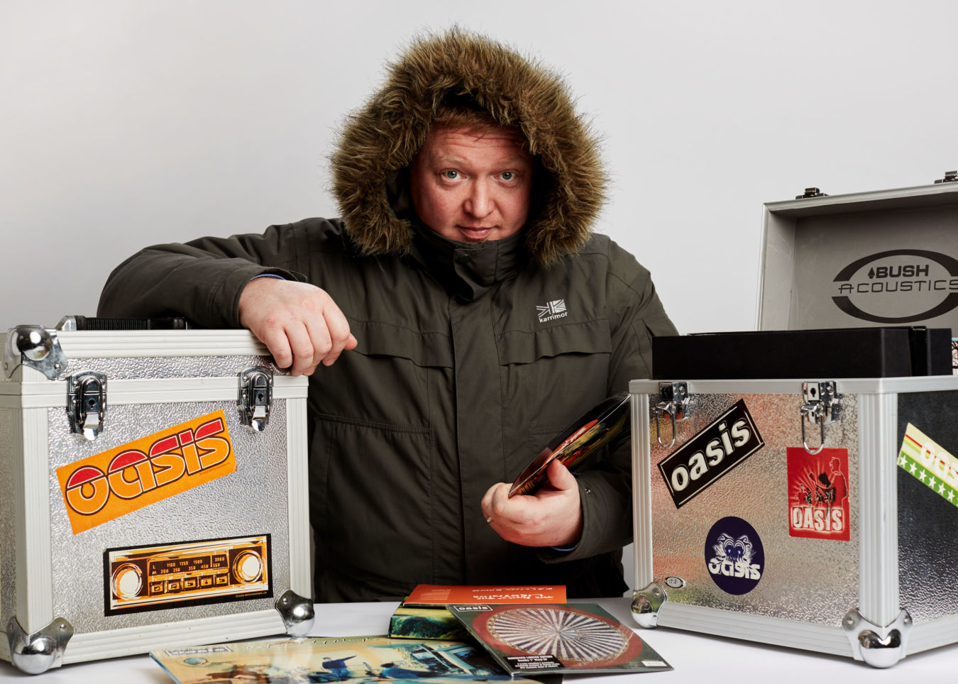 A person in a fur-lined coat holds a vinyl record beside two metal cases decorated with Oasis stickers. One case is open, revealing "BUSH ACOUSTICS" text inside, surrounded by scattered albums.