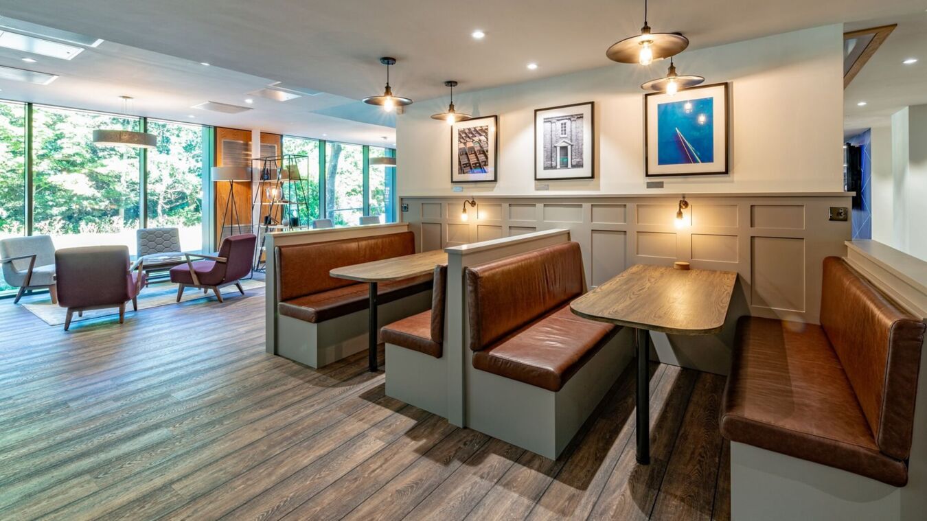 Booth seating with wooden tables and brown leather cushions is arranged across from large windows. Framed art decorates the cream-paneled walls, and cozy chairs sit in a lounge area.