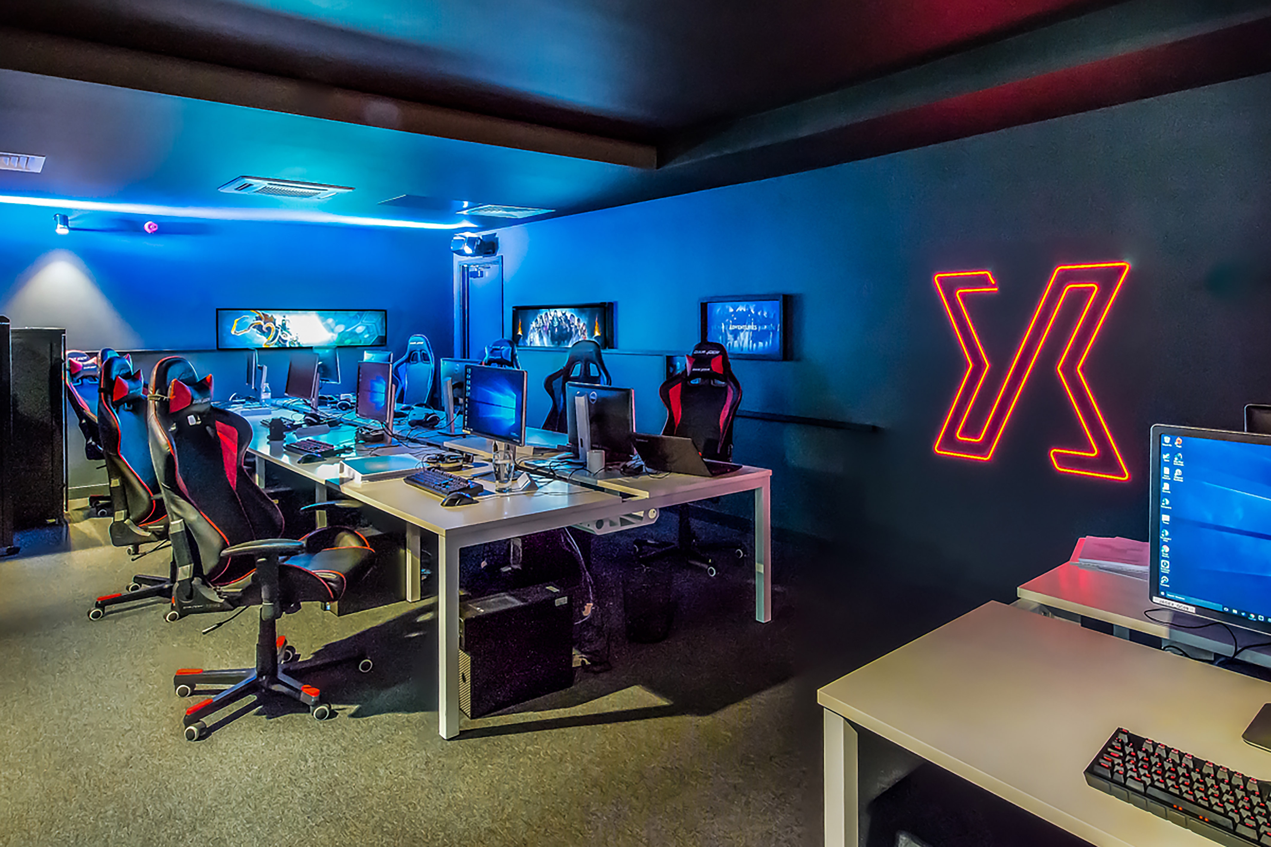 Gaming desktops with monitors and chairs are arranged in a dimly lit room, featuring a glowing red logo on a blue wall, and multiple screens displaying blue backgrounds.
