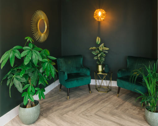 Cozy room with dark green walls, two green velvet chairs, a potted plant, gold geometric light fixture, mirror, and wooden herringbone floor. Warm and inviting.