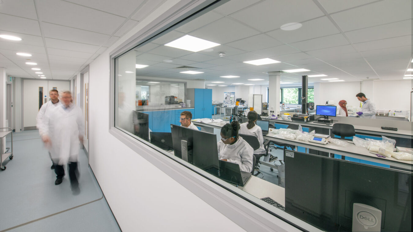 View through an interior window into a busy office or laboratory, where employees are working at desks with computers under bright fluorescent lighting.