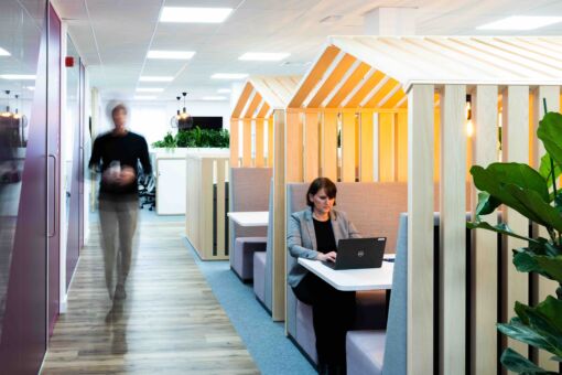 Modern office space with wooden-framed booth seating resembling small house structures, where a person is working on a laptop under warm lighting