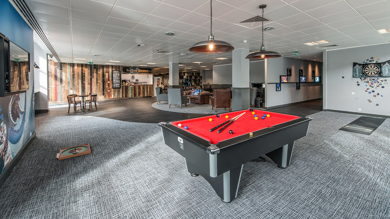A red pool table with balls and cues stands in a modern lounge featuring wooden walls, a bar labeled “Golden Gnome,” dartboard area, and seating with chairs and tables.