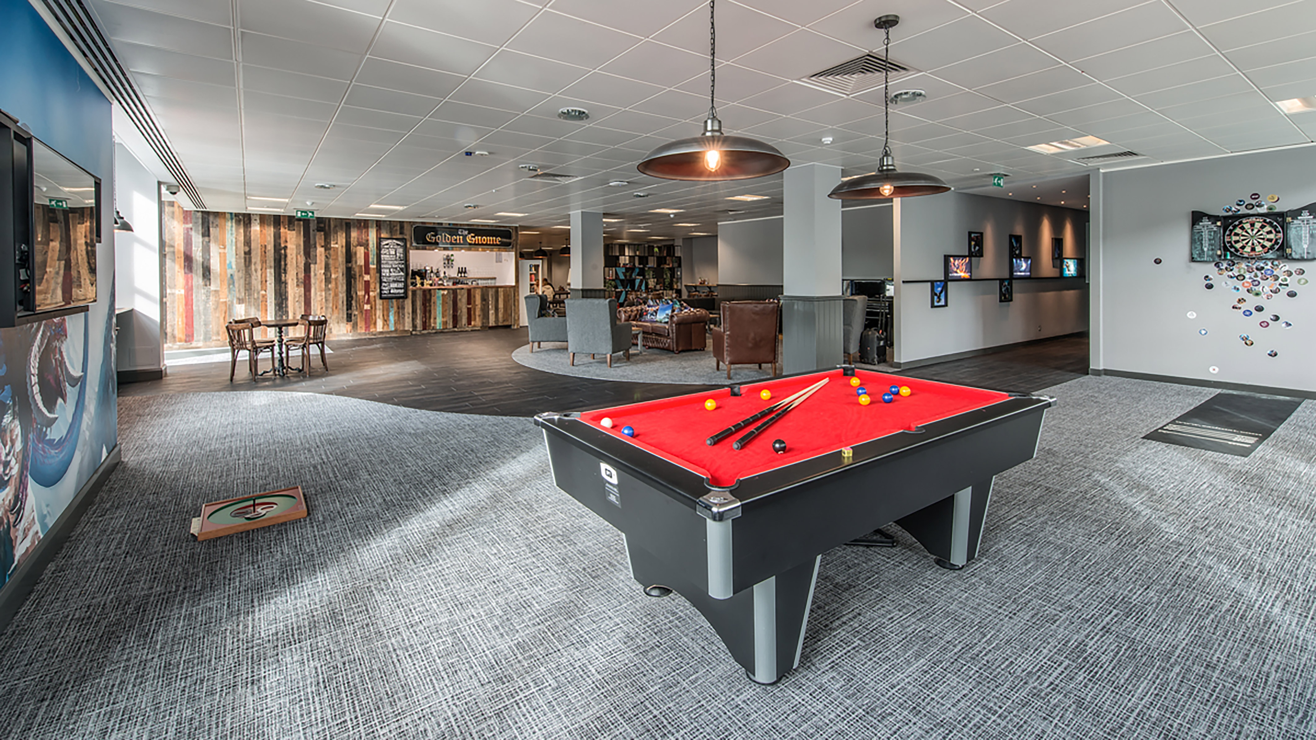 A red pool table with balls and cues stands in a modern lounge featuring wooden walls, a bar labeled “Golden Gnome,” dartboard area, and seating with chairs and tables.