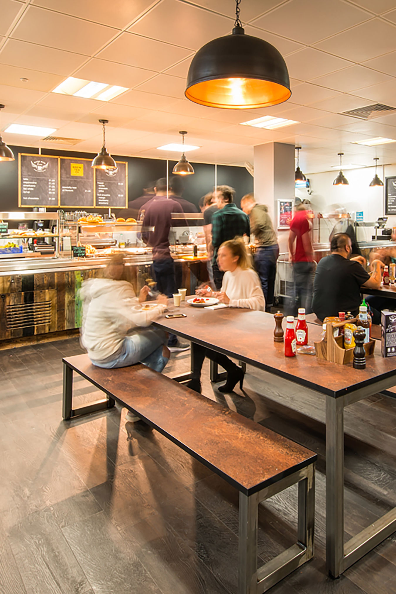 Cafeteria tables filled with people eating and conversing, in a bright, modern setting with overhead lighting. A food counter in the background offers various meal options.