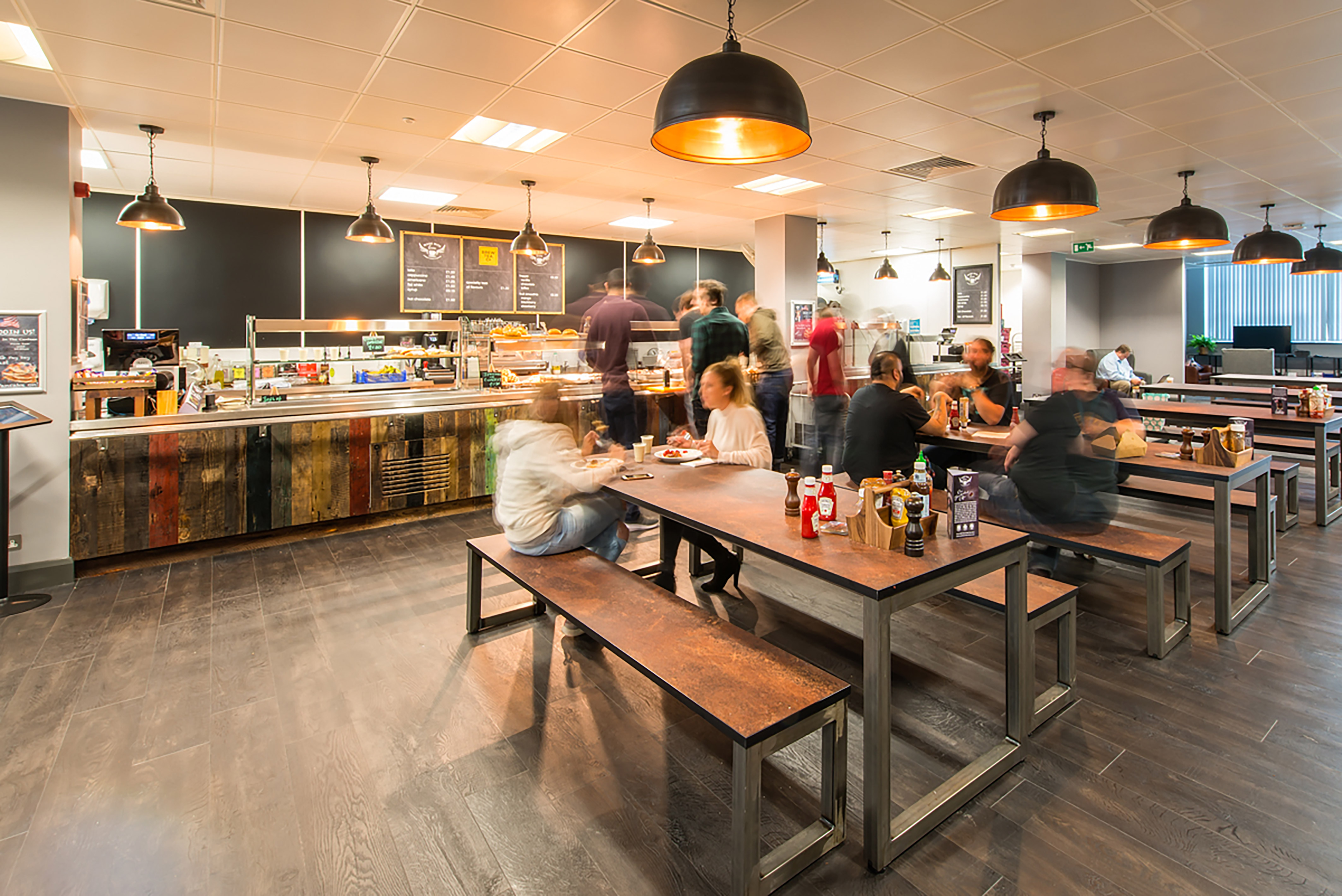 Cafeteria tables filled with people eating and conversing, in a bright, modern setting with overhead lighting. A food counter in the background offers various meal options.