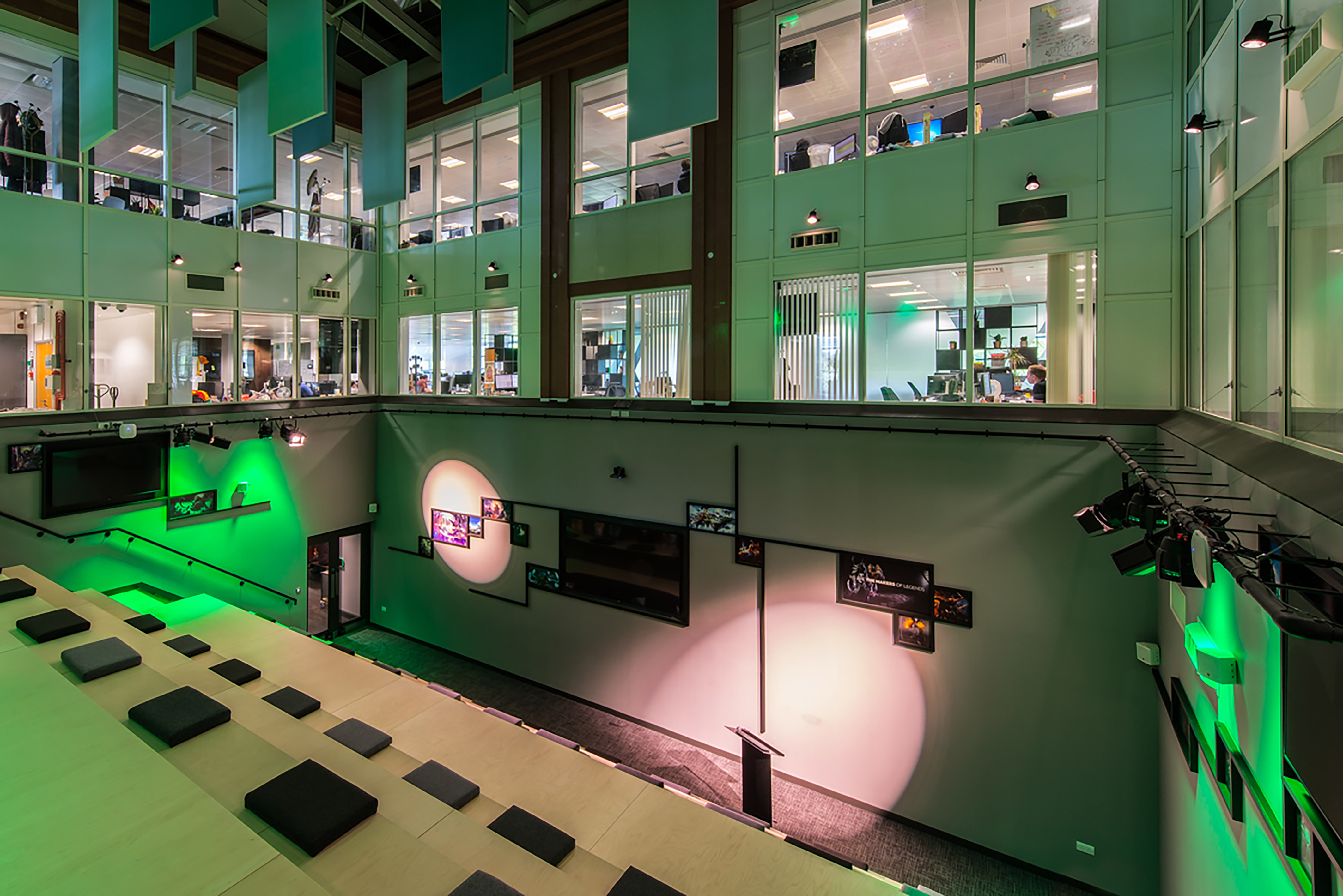 An open office atrium features tiered seating with cushions, illuminated by green lighting, surrounded by glass-walled offices above. The wall displays photos and is highlighted by spotlights.