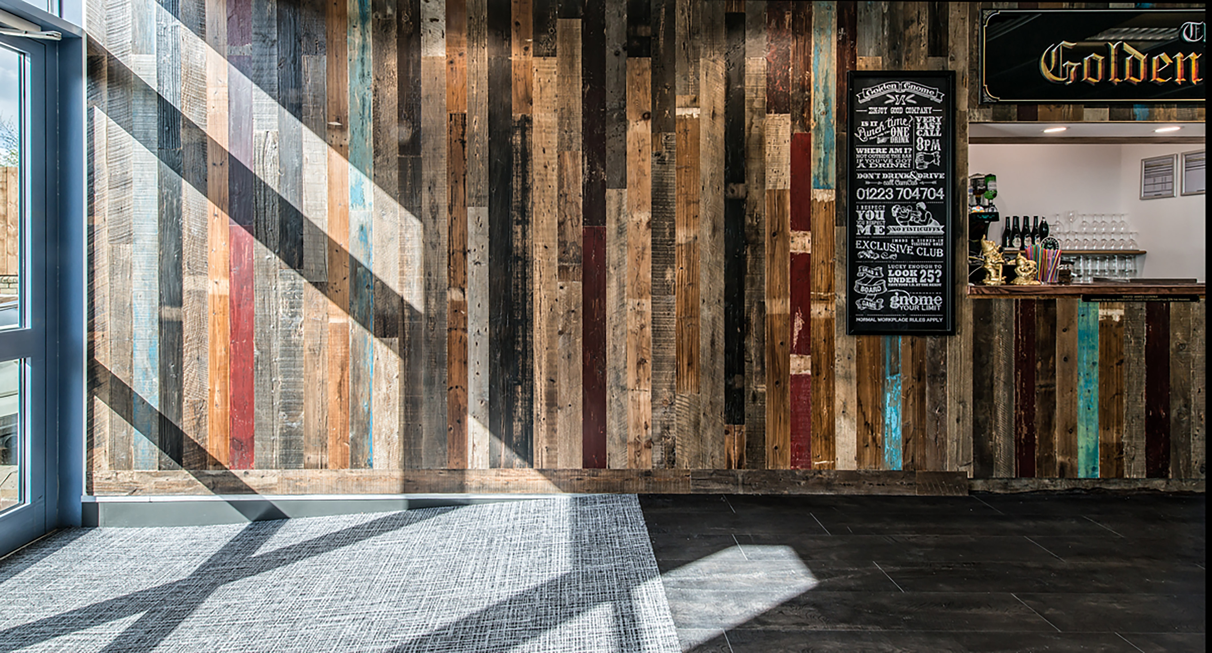 Wooden wall paneling with colorful vertical planks forms the backdrop for a bar area; chalkboard signage reads "Golden Chrome," listing club rules, under a sign with partial text "Golden." Sunlight creates shadow patterns across the wall and carpeted floor.