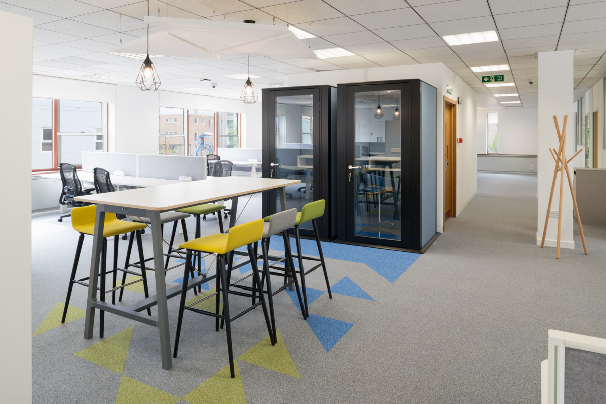 A modern office features high tables with chairs, geometric-patterned carpet, glass meeting booths, and ceiling lights, creating a bright, collaborative workspace with large windows overlooking buildings.