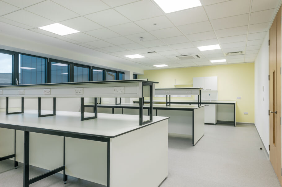 Bright laboratory room with white workbenches, black framing, overhead shelving, tiled flooring, and fluorescent ceiling lighting.