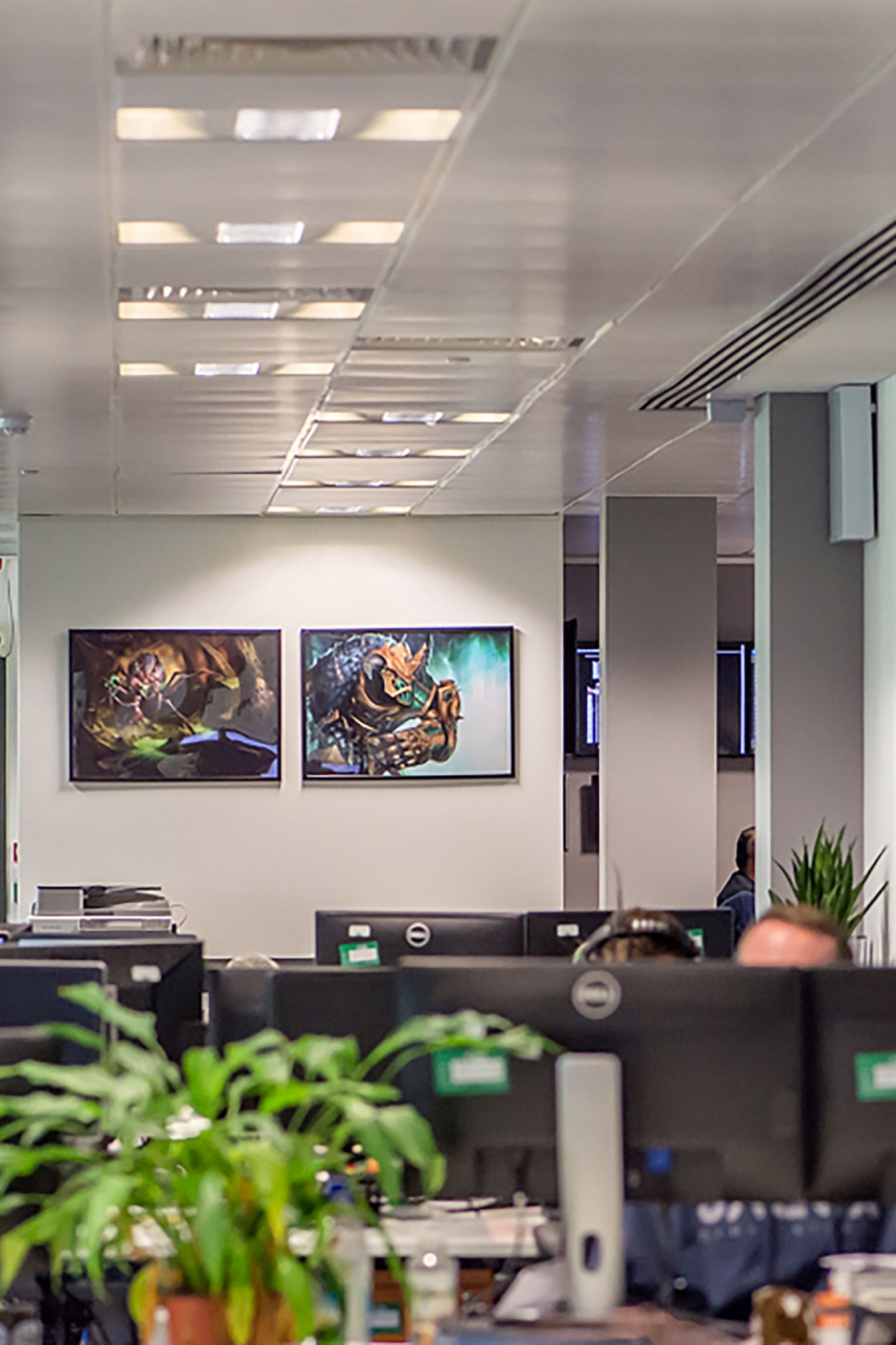 Office setting featuring computer monitors and a green plant in the foreground, with two fantasy-themed paintings on a wall in the background. The ceiling has recessed lighting.