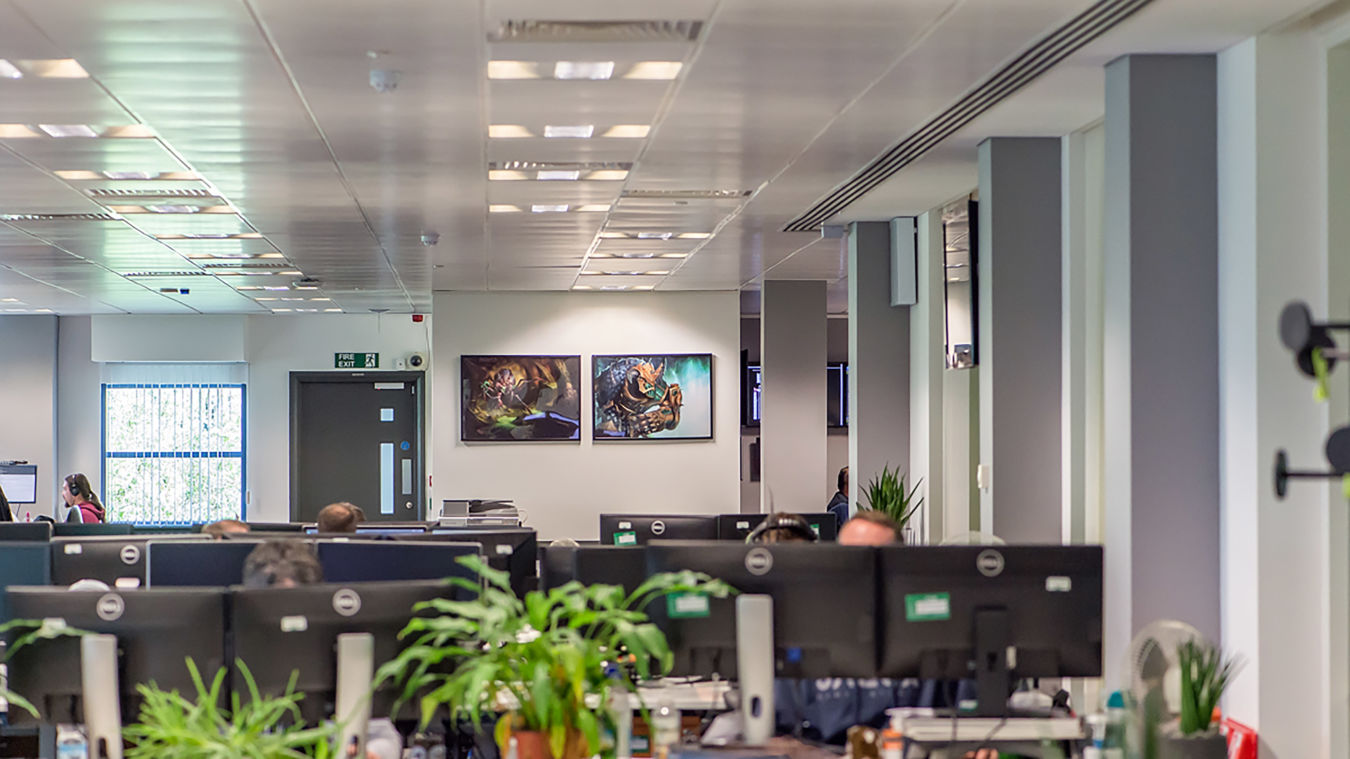 Office setting featuring computer monitors and a green plant in the foreground, with two fantasy-themed paintings on a wall in the background. The ceiling has recessed lighting.