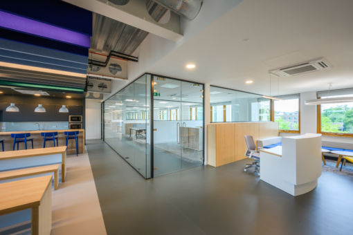 A bright, contemporary office space features a white reception desk with a blue chair, a glass-walled office area, and a kitchen/break area with dark cabinetry, light wooden tables, and blue bar stools. Natural light streams in from large windows on the right, offering a glimpse of greenery outside.
