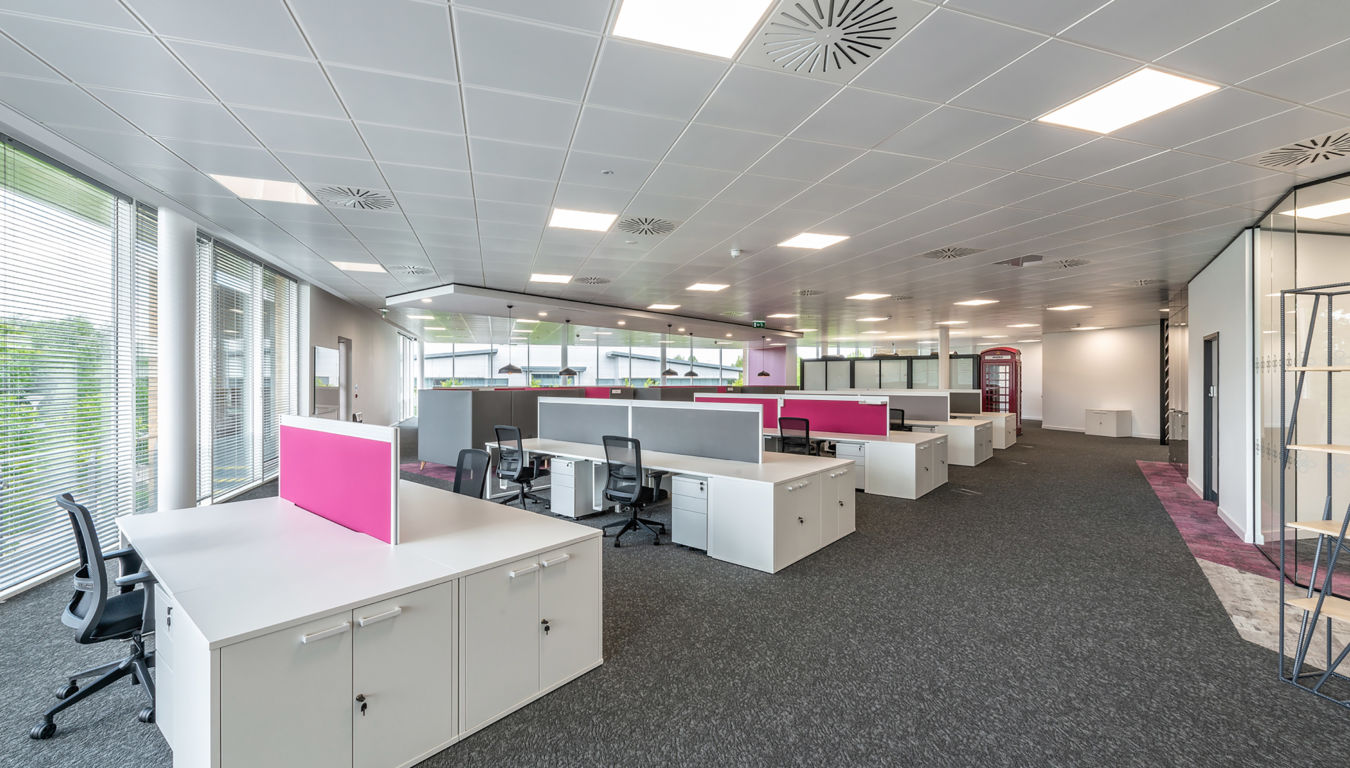 Modern open office space with white desks, pink dividers, and gray chairs. Large windows on the left let in natural light, creating a bright, spacious atmosphere.