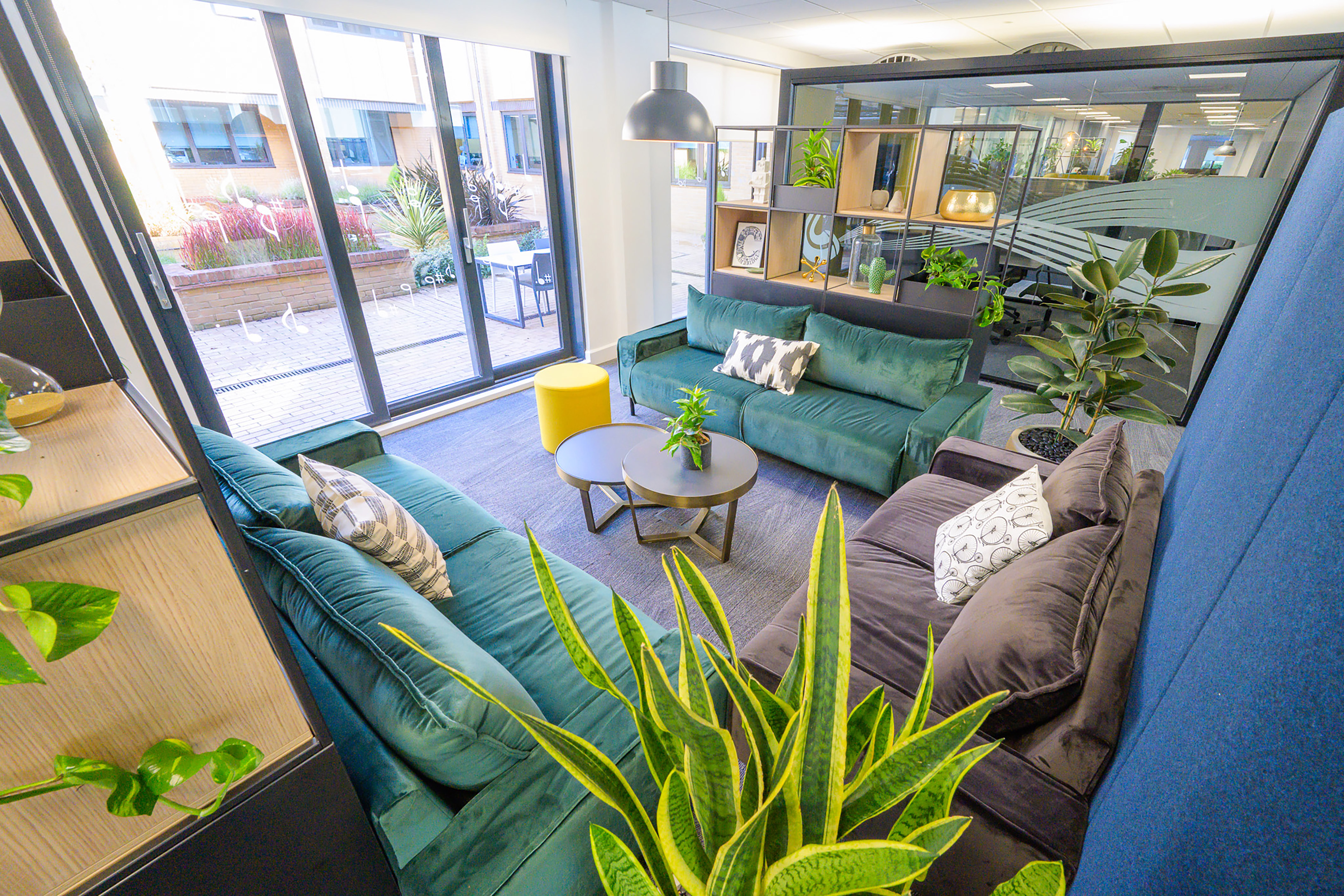 Contemporary office lounge featuring green sofas surrounding a round table with a plant; large windows offer a view of an outdoor area with plants and seating.