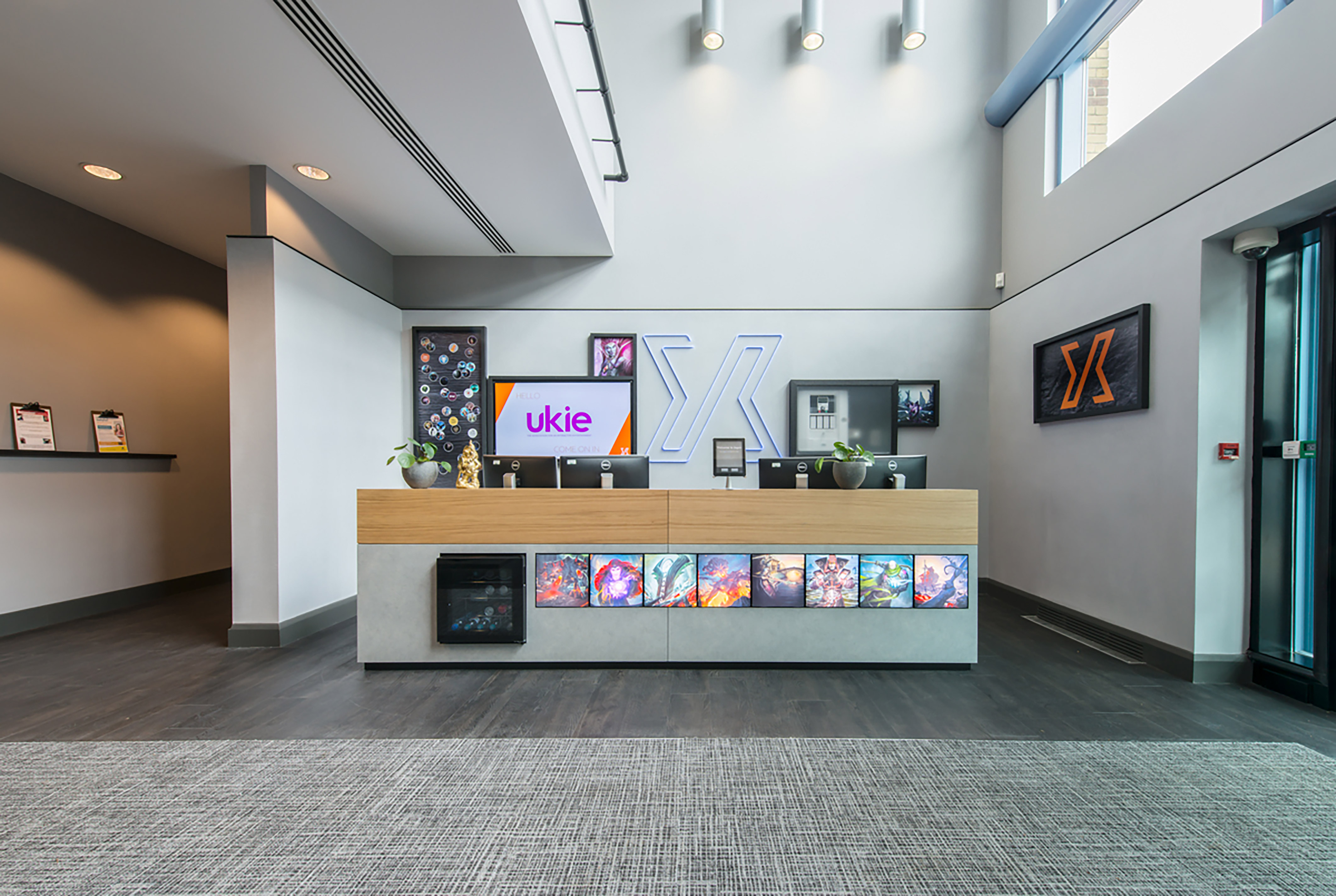 Modern reception desk displaying colorful video game artwork, with a monitor showing "Hello ukie" in a bright, airy office environment featuring potted plants and wall logos.