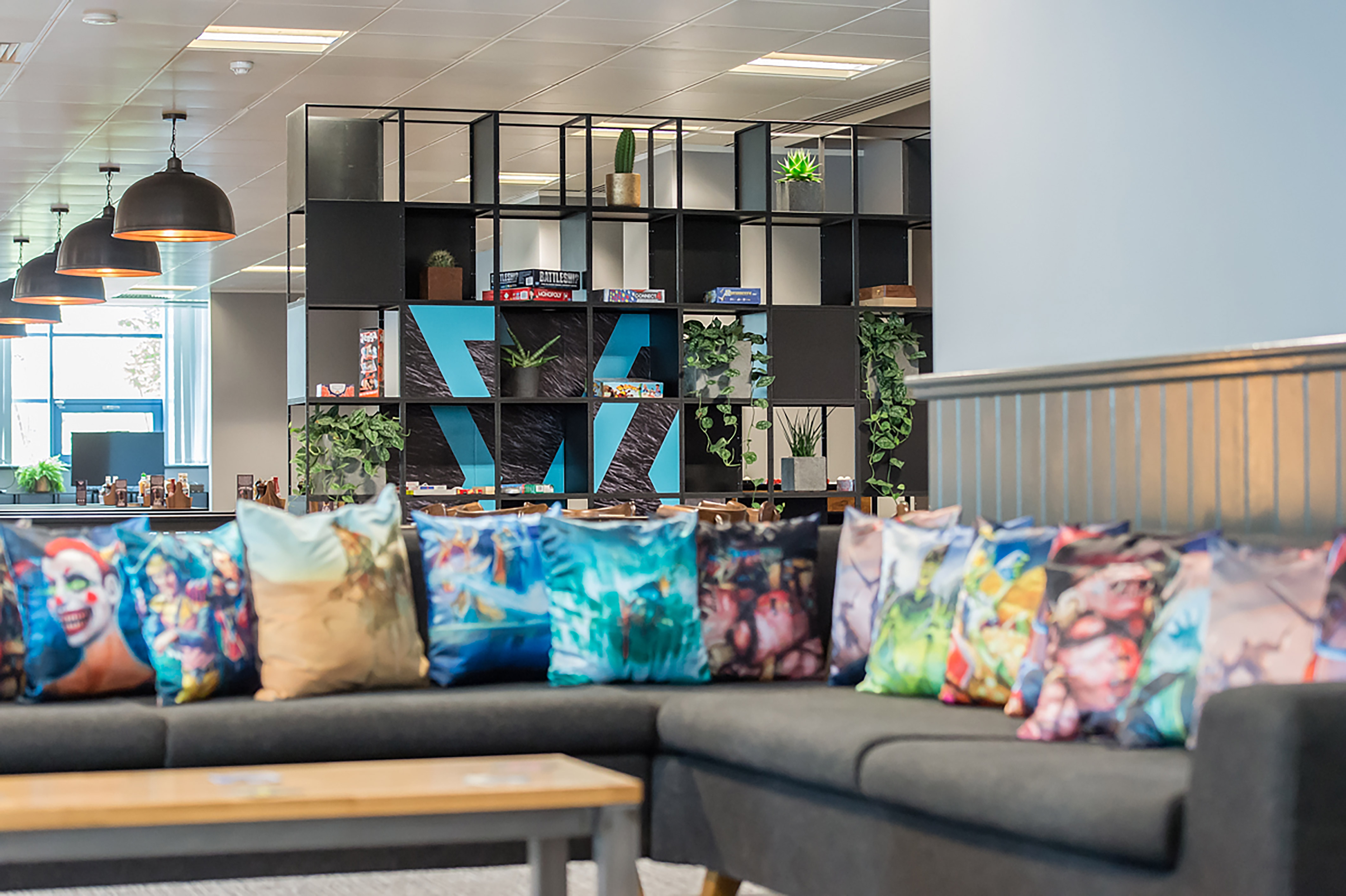 Modern office lounge with a gray couch filled with colorful graphic pillows. In the background, a black shelving unit holds plants and board games, creating a playful, relaxed atmosphere