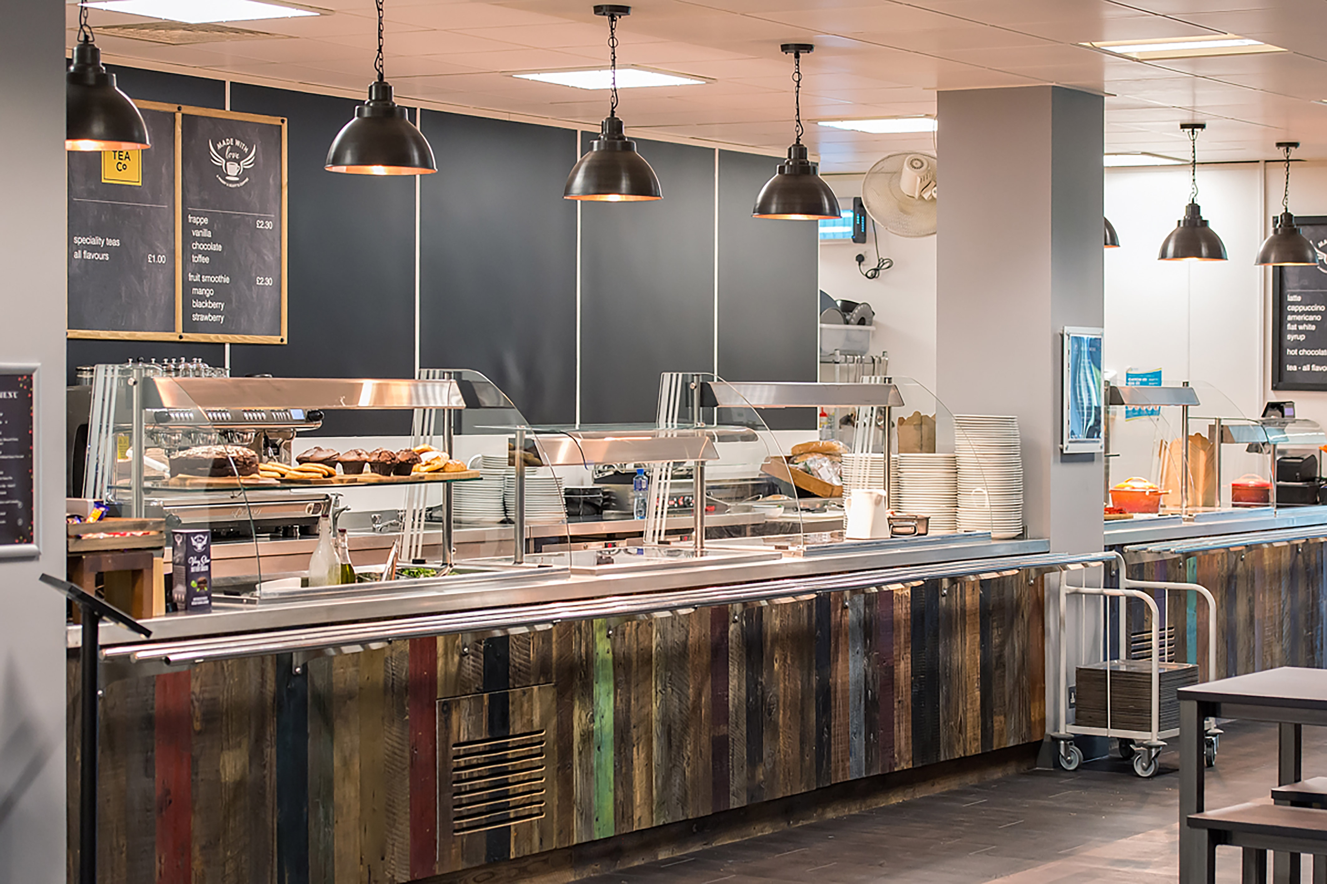 Cafeteria counter displaying various baked goods under glass covers, surrounded by industrial-style decor with pendant lights. Wall menus include “TEA Co” and drink prices like “frappe” at £2.30.