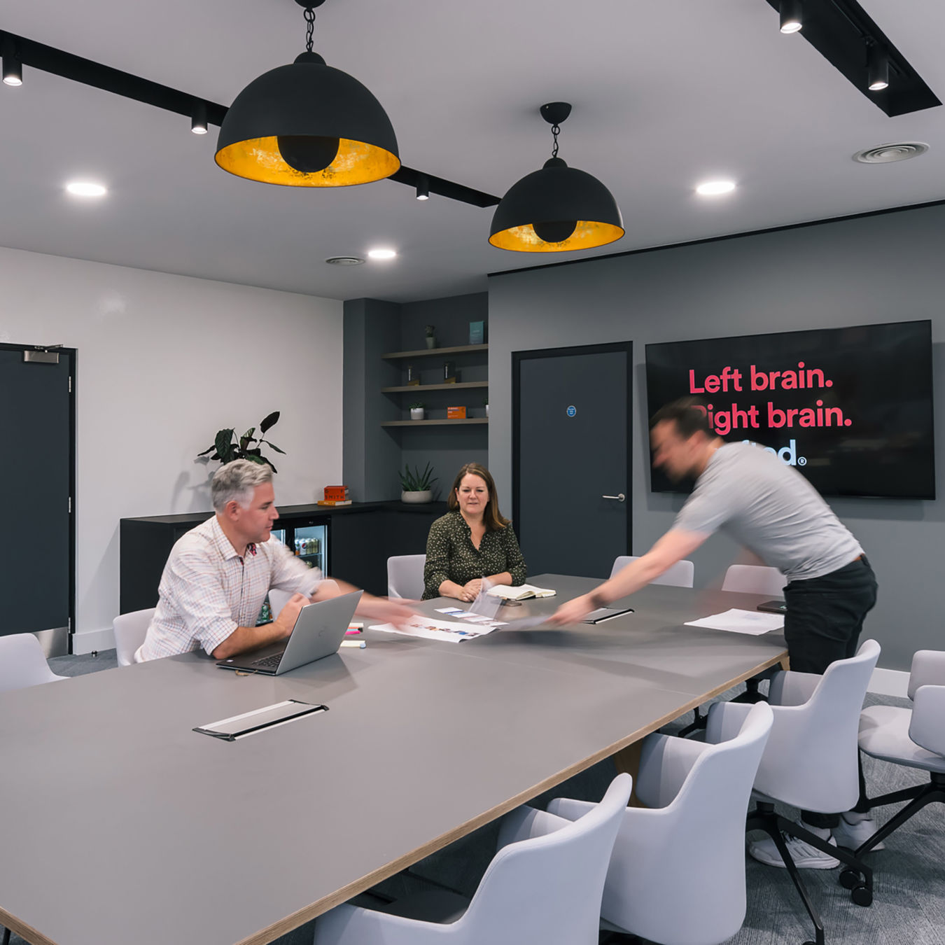 Modern conference room with three people around a large table. One is using a laptop, another is passing documents.