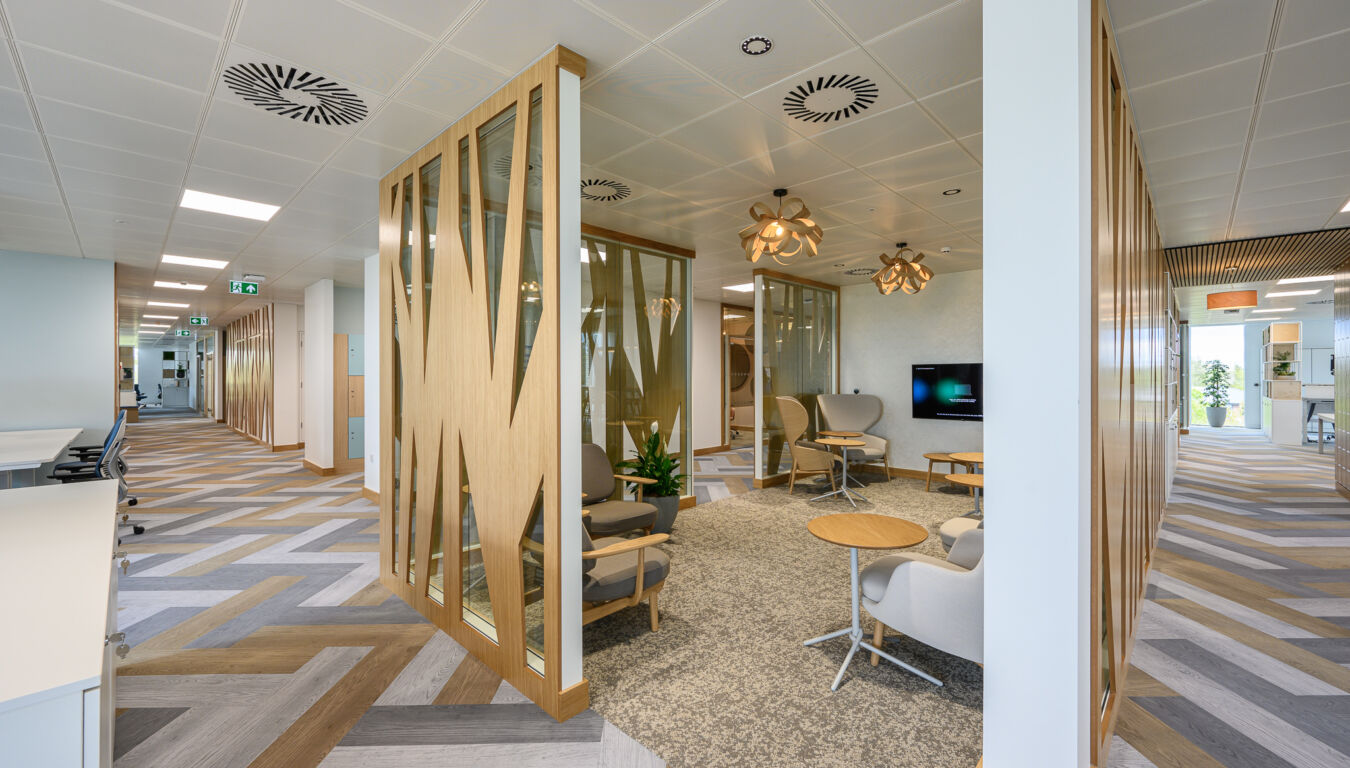 Modern office interior with wood accents and glass walls, featuring cozy seating, contemporary pendant lights, patterned flooring, and a TV. Bright and inviting