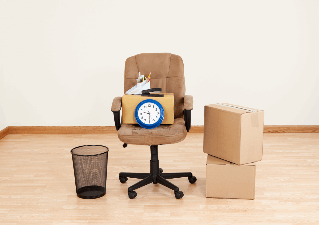 Office chair with a cardboard box containing office supplies and a clock, surrounded by moving boxes and a wastebasket, symbolizing relocation or job transition