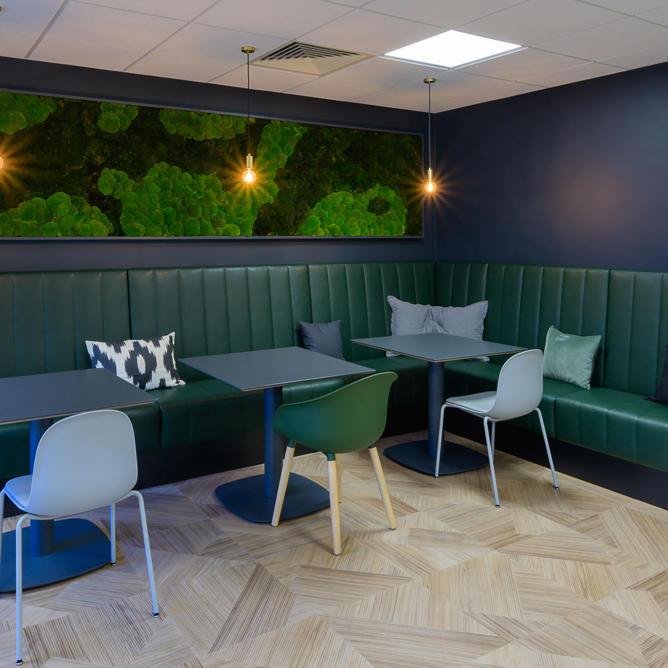 Modern seating area with green leather banquette against navy walls. Features minimalist tables, various colored chairs, and warm pendant lights.
