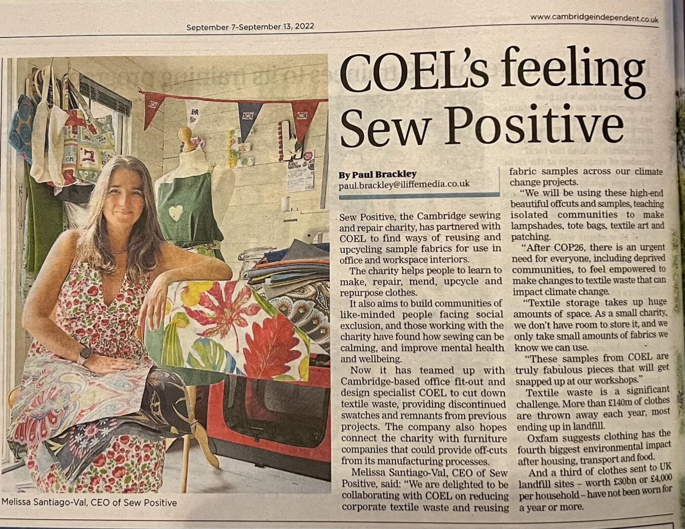 A woman sits in a room filled with colorful fabrics and bags, alongside a mannequin. Text heading reads “COEL’s feeling Sew Positive". Article by Paul Brackley discusses a sewing charity partnership.