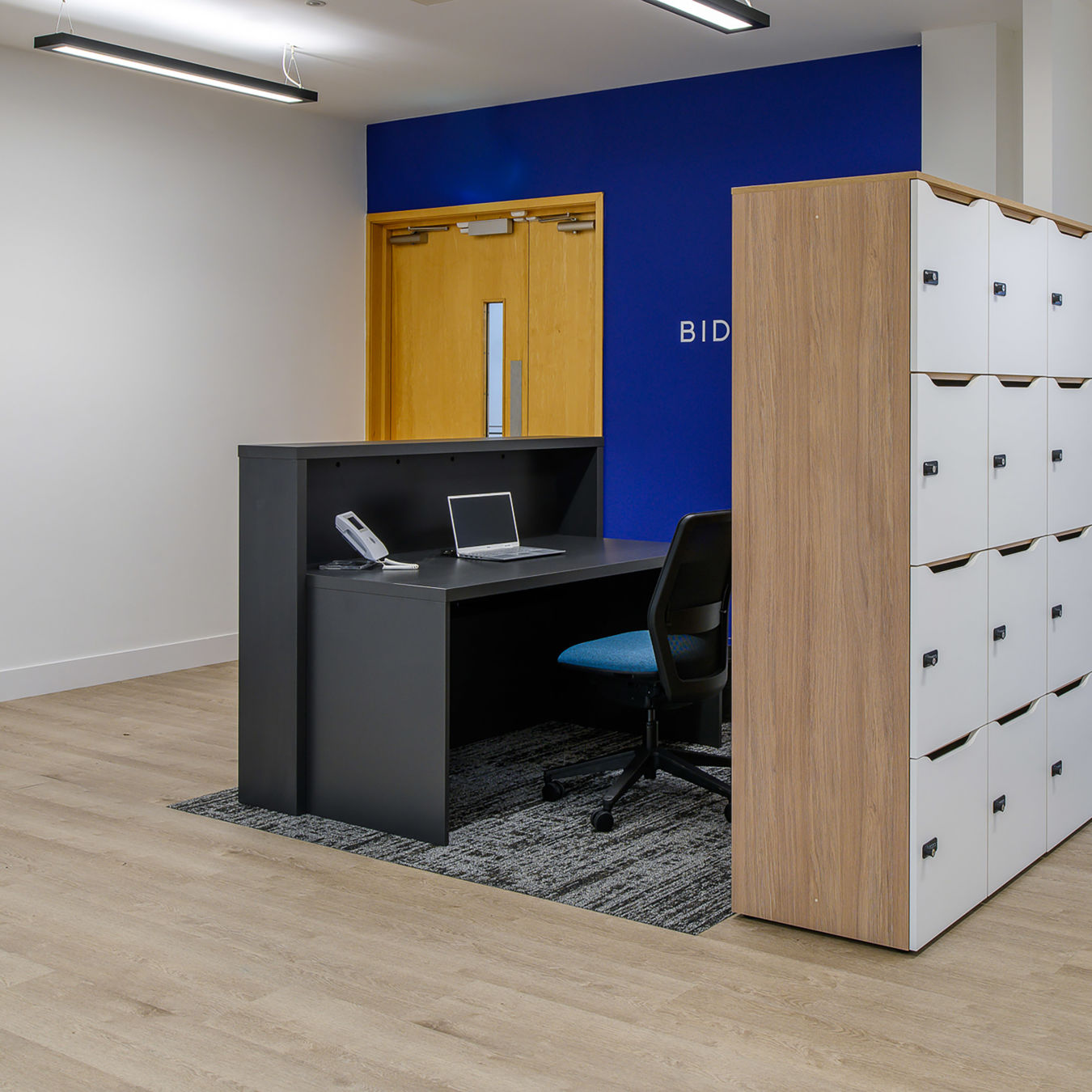 Modern office reception area with a wooden desk, laptop, phone, and an empty chair. White lockers line one side, contrasting with a blue accent wall