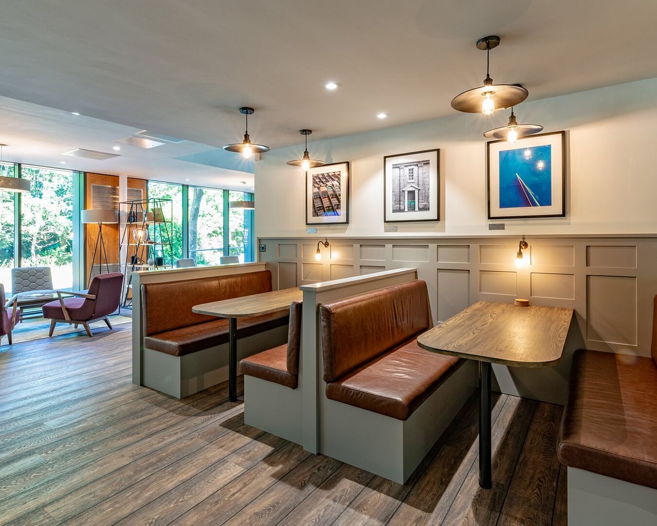 Booths with brown leather seats and wooden tables are lined against a white-paneled wall, decorated with framed photographs. Large windows reveal greenery, while modern chairs occupy adjacent open space.