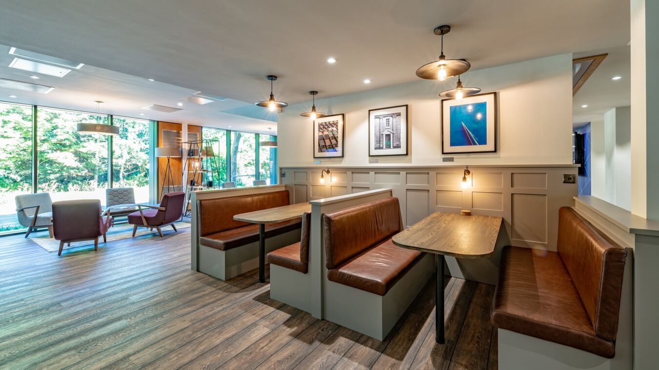 Booths with brown leather seats and wooden tables are lined against a white-paneled wall, decorated with framed photographs. Large windows reveal greenery, while modern chairs occupy adjacent open space.