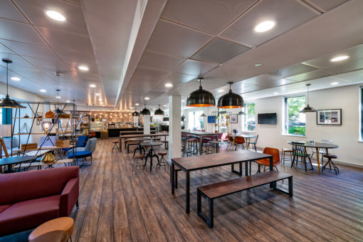Spacious and modern cafeteria or communal dining area featuring wooden floors, various seating options, pendant lighting, and a well-lit open layout