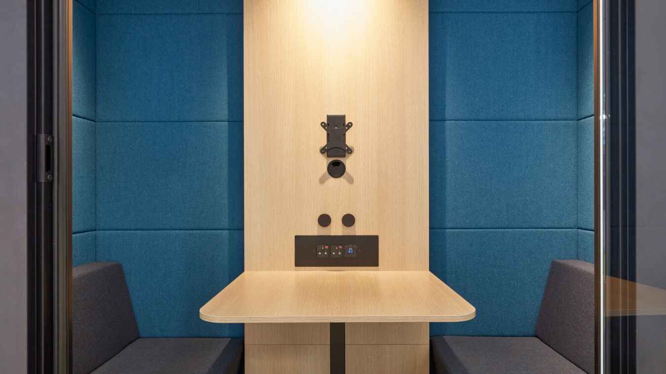 A modern, minimalistic booth features a wooden table, wall-mounted equipment, and two cushioned benches. The blue upholstered walls create a cozy, enclosed space ideal for individual work or meetings.