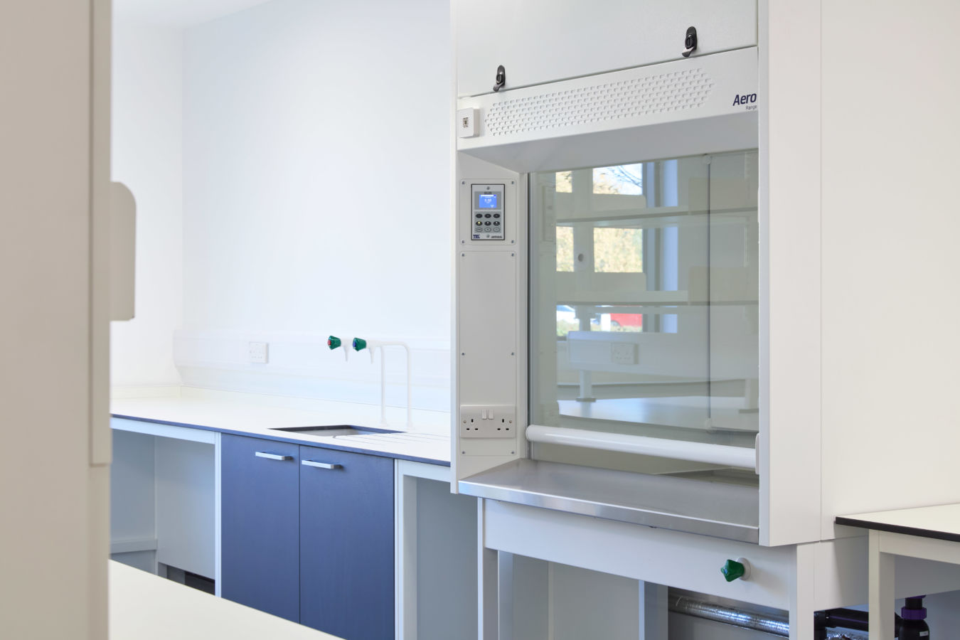 Modern laboratory with blue cabinets and a white fume hood on the right. Bright lighting and clean surfaces create a sterile, professional atmosphere