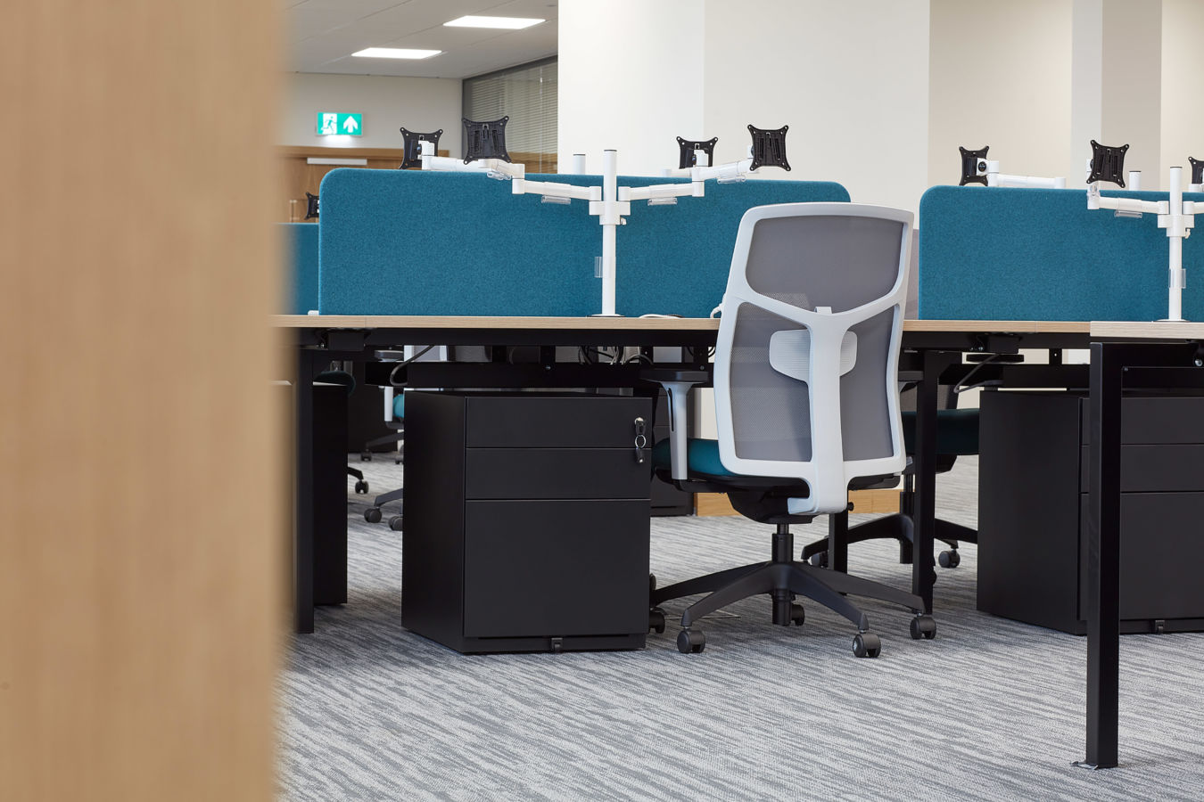 close up image of a single person desk, with blue divider panel a white and blue chair and a filling cabinet under the desk