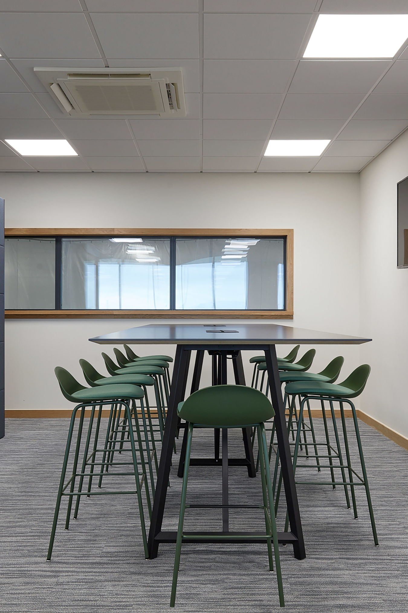 Modern office meeting space with a long table and six green chairs. A flat-screen TV is on the wall, and large windows provide natural light.