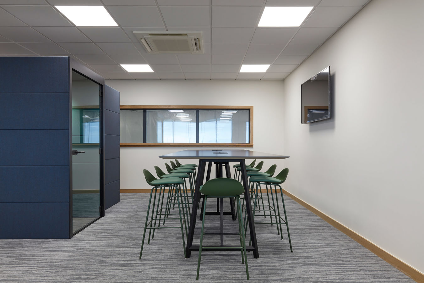 Modern office meeting space with a long table and six green chairs. A flat-screen TV is on the wall, and large windows provide natural light.